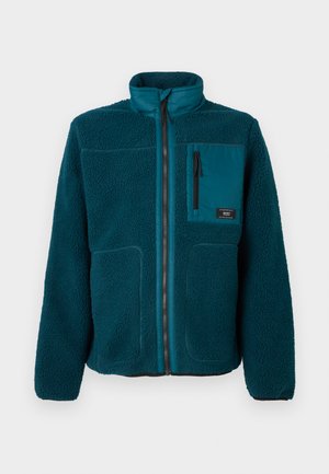 Veste en polaire bleu sarcelle avec zip sur toute la longueur, col montant, poche poitrine zippée et deux poches inférieures à fente.