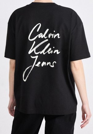 Osoba v černém tričku s krátkými rukávy a bílým nápisem "Calvin Klein Jeans" na zádech, stojící před jednobarevným pozadím.