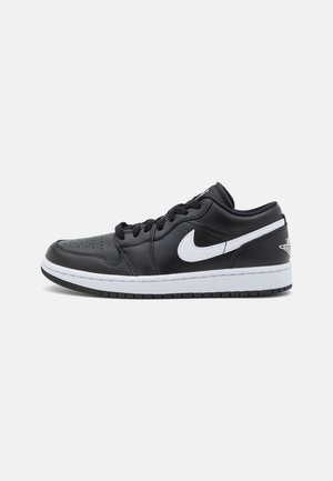 Baskets basses noires avec un swoosh Nike blanc, une semelle intermédiaire blanche, une semelle extérieure noire et des lacets sur un fond gris clair uni.