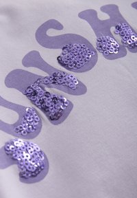 Vitt tyg med lila paljetter ordnade i ett stiliserat textmönster. Paljetterna varierar i storlek och reflekterar ljus, vilket ger textur.