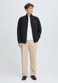 Veste noire zippée avec col haut, chemise blanche en dessous, pantalon beige et chaussures noires. Design simple avec logo sur la poitrine.