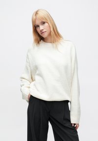 Kremowy, oversize'owy sweter z zaokrąglonym dekoltem i ściągaczami na rękawach, w połączeniu z ciemnymi plisowanymi spodniami. Miękka tekstura, luźny krój.