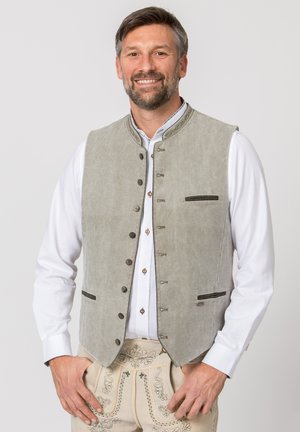 Lächelnder Mann mit hellgrauer traditioneller Weste über weißem Hemd und bestickten beigen Lederhosen, die Hände ruhen in der Nähe der Taschen.