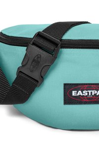 Teal tygmidjeväska med svart justerbart rem och spänne, med en svart logotypfynd med röd text. Dragkedja synlig.