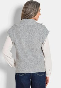 Grauer ärmelloser Pullover mit ribbeliger Textur, weitem Kragen und leicht verkürztem hinterem Saum, schichtweise über einem weißen Langarmshirt getragen.