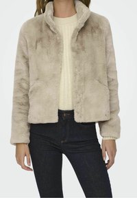 Fuskpälsjacka i ljusbeige med sjalkrage, långa ärmar och två framfickor, ihop med en krämfärgad ribbvävd tröja och mörka jeans.