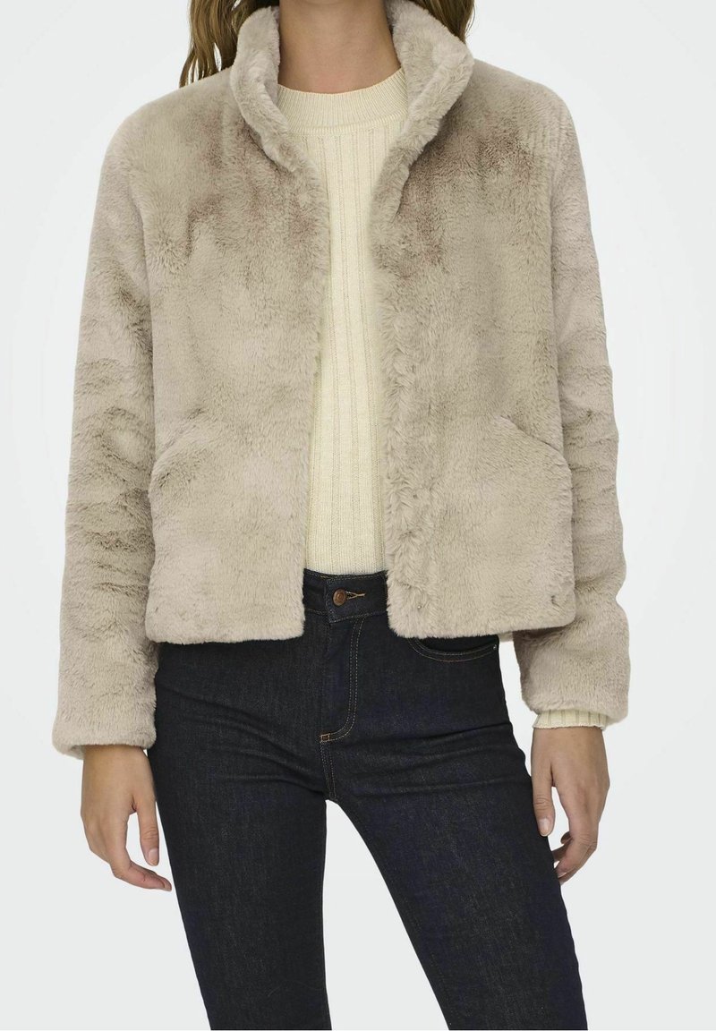 Fuskpälsjacka i ljusbeige med sjalkrage, långa ärmar och två framfickor, ihop med en krämfärgad ribbvävd tröja och mörka jeans.