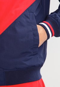 Marinblå vindjacka med röda diagonala detaljer, med en sidoficka och ribbade muddarna med röd, vit och marinblå rand.