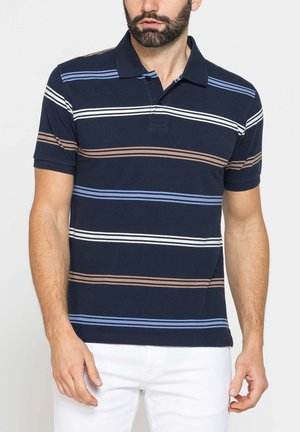 Uomo che indossa una polo blu navy con strisce orizzontali bianche, blu e marroni, abbinata a pantaloni bianchi, in piedi su uno sfondo chiaro.