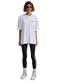 Fehér, túlméretezett póló kék logóval, fekete leggingssel, amelyen fehér és kék csíkok vannak, valamint fehér edzőcipő zöld részletekkel.