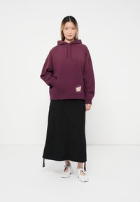 Felpa con cappuccio bordeaux con tasca frontale e logo, abbinata a una gonna lunga nera. Modella che indossa scarpe grigie dalla forma unica.
