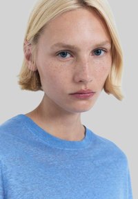 Jasnoniebieski sweter dzianinowy z okrągłym dekoltem, miękką teksturą i delikatnym melanżowym wzorem. Modelka ma jasnoblond włosy i naturalny makijaż.