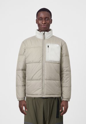 Mann trägt eine hellbeige gesteppte Jacke mit hohem Kragen und Reißverschlusstasche an der Brust, steht vor einem schlichten weißen Hintergrund.