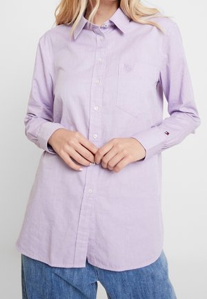 Frau trägt ein langärmliges, hellviolettes Hemd mit Brusttasche und kleinem Logo auf der Manschette, kombiniert mit blauen Jeans.