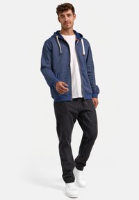 Sudadera azul con cremallera y un patrón texturizado, capucha con cordón, bolsillos frontales, combinada con jeans oscuros y zapatillas blancas.