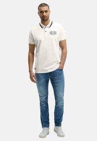 Man in een off-white polo met tekst en blauwe spijkerbroek, staand met één hand in zijn zak en witte sneakers op een grijze achtergrond.