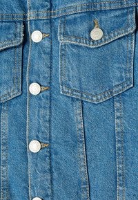 Knappad denimjacka i ljusblått tyg med dubbla bröstfickor, silverknappar och kontrasterande gula sömmar.