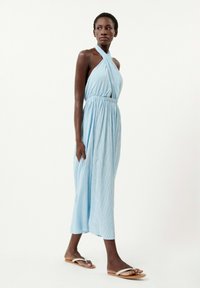 Hellblaues Maxikleid mit Trägern und vertikalen Streifen, elastischem Bund und fließendem Schnitt. Kombiniert mit flachen Sandalen mit minimalen Riemen.