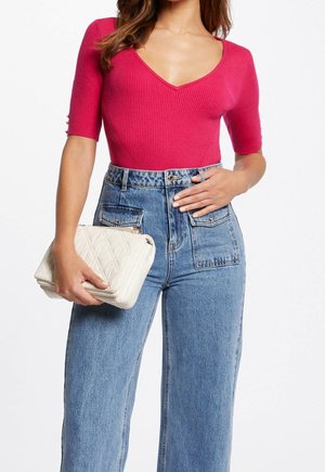 Femme portant un haut fuchsia ajusté à manches courtes et un jean bleu taille haute, tenant une pochette blanche matelassée à hauteur de taille.