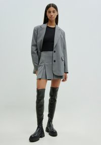 Grauer oversized Blazer mit zwei Knöpfen, einem eng anliegenden schwarzen Oberteil, einem gefalteten Minirock und kniehohen schwarzen Stiefeln mit klobigen Sohlen.