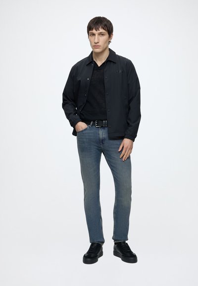 Jeune homme debout, portant une veste noire, une chemise noire, un jean bleu, une ceinture noire et des chaussures noires sur fond blanc uni.