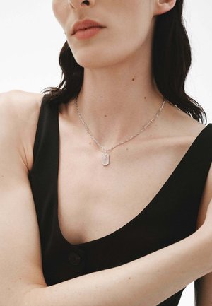 Collar de cadena de plata con un colgante rectangular grabado en rojo con la palabra "SURGE". La modelo lleva una blusa negra con un escote pronunciado.