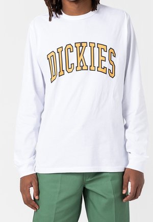 Persona con camisa blanca de manga larga con texto amarillo grande y arqueado "DICKIES" en el pecho y pantalones verdes, de pie contra un fondo liso.
