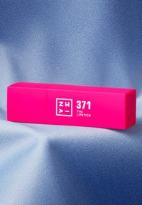Lys rosa rektangulær leppestiftetui merket "371 THE LIPSTICK," med en glatt matt finish, plassert på et mykt blått tekstilbakgrunn.