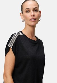 Čierny top s krátkym rukávom a okrúhlym výstrihom, so zdobnou čipkovou detailou na pleciach, vyrobený z mäkkej, ľahkej látky.