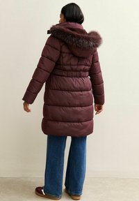 Parka acolchada color burdeos con capucha de pelo, cintura ceñida con cinturón y mangas largas, combinada con jeans de pierna ancha y calzado marrón.
