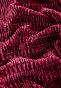 Tiefrotes, strukturiertes Gewebe mit erhobenen, wellenförmigen Mustern. Das Material wirkt weich und plüschig und zeigt unterschiedliche Lichtreflexionen.