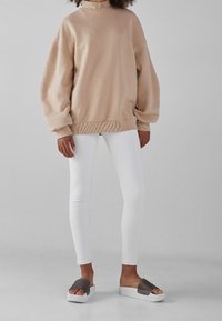 Beige oversize-tröja med ribbad fåll, kombinerad med vita tajta jeans och gråa slip-in sandaler med tjock vit sula. Mjuk tygstruktur.