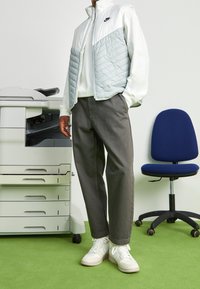 Ljusgrå och vit puffjacka med dragkedja, tillsammans med mörkgrå byxor och vita sneakers. Bakgrunden visar en skrivare och en grön matta.