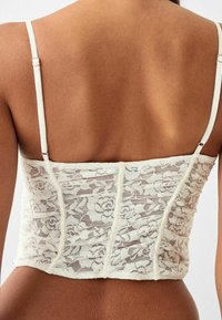 Krajkový korzetový top s nastavitelnými ramínky, zdobený květinovým vzorem, s nařasenou strukturou a světlou krémovou barvou.