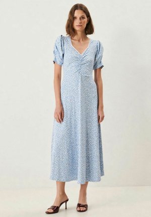 Nainen pukeutuneena vaaleansiniseen midimekkoon, jossa on liehuvat hihat ja hienovarainen kukkakuosi, yhdistettynä tummiin korkokenkiin, seisomassa yksiväristä taustaa vasten.