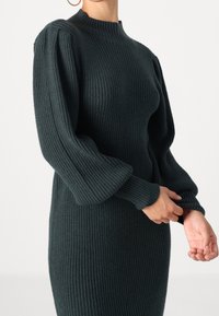 Abito lavorato a maglia in verde scuro, con collo alto e maniche a sbuffo. Presenta un corpetto aderente e maniche ampie, creando un effetto tridimensionale.