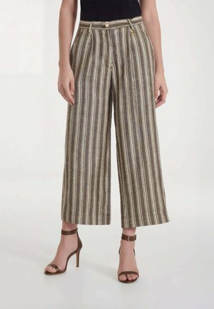 Donna che indossa pantaloni a righe a vita alta e gamba larga abbinati a sandali marroni con tacco alto e top nero, in piedi davanti a uno sfondo semplice.