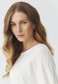 Hvid sweater med ribbet tekstur og bred halsudskæring, parret med gyldne hoops øreringe. Lange, bølgede hair indrammer ansigtet. Blødt lys.