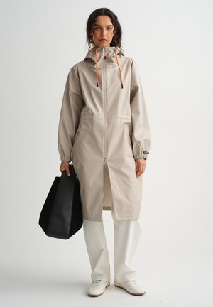 Nainen pukeutuneena beigeen hupulliseen sadetakkiin, jossa oranssit kiristysnauhat, valkoiset housut, valkoiset tennarit ja hän kantaa suurta mustaa kangaskassia.