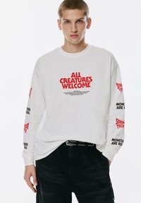 Junger Mann trägt ein weißes Langarmshirt mit rotem und schwarzem Text: "ALLE KREATUREN WILLKOMMEN" auf der Brust, "STRANGER THINGS" und "MONSTER SIND REAL" auf den Ärmeln.