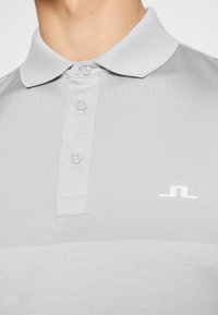 Ljusgrå polo med tre knappar och en liten vit geometrisk logotyp på bröstet, bärs av en person.