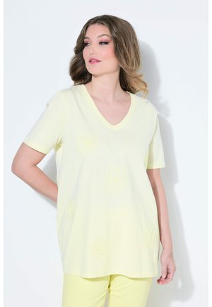 Frau mit welligem Haar, die ein hellgelbes V-Ausschnitt-Kurzarm-Oberteil und passende Hose trägt, stehend vor einem einfachen weißen Hintergrund.