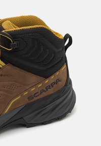 Scarpa pārgājienu zābaki ar brūnu zīda ķermeni, melnu auduma apkakli un dzelteniem akcentiem. Iekļauj teksturētu augšpusi un izturīgu gumijas zoli.