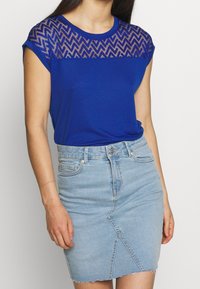 Camiseta azul con yugo de malla texturizada de patrón en forma de chevron y mangas cortas, combinada con una falda de mezclilla azul clara ajustada con dobladillo deshilachado.