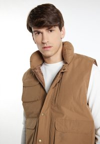 Braun gepolsterte Weste mit hohem Kragen, mehreren Taschen und einem Reißverschluss. Der Stoff wirkt glatt mit mattem Finish und Druckknöpfen.