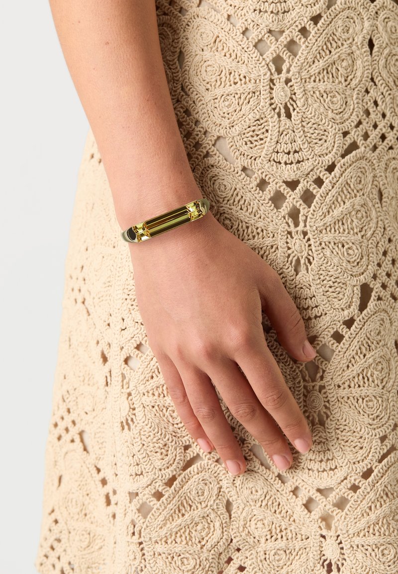 Main portant un bracelet en or avec des pierres précieuses rectangulaires reposant sur un tissu beige crocheté avec des motifs de papillons.