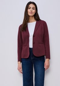 Femme aux longs cheveux bruns portant un blazer bordeaux, une chemise blanche et un jean bleu foncé, debout devant un fond neutre.