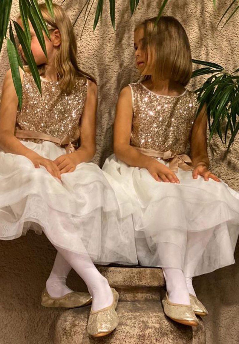 Zwei Mädchen sitzen nebeneinander in Kleidern mit goldenen Pailletten-Oberteilen und weißen, schichten Tüllröcken. Sie tragen weiße Strumpfhosen und goldene Schuhe.