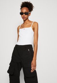 Hvit spaghetti-stropp bodysuit med en liten rød logo, kombinert med svarte cargo-bukser med glidelåser og sidelommer. Glatt, tettsittende stoff.