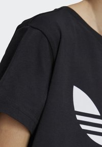 T-shirt noir en coton avec un design de logo blanc comportant trois rayures et une forme de feuille. Encolure en V et légère texture visible dans le tissu.